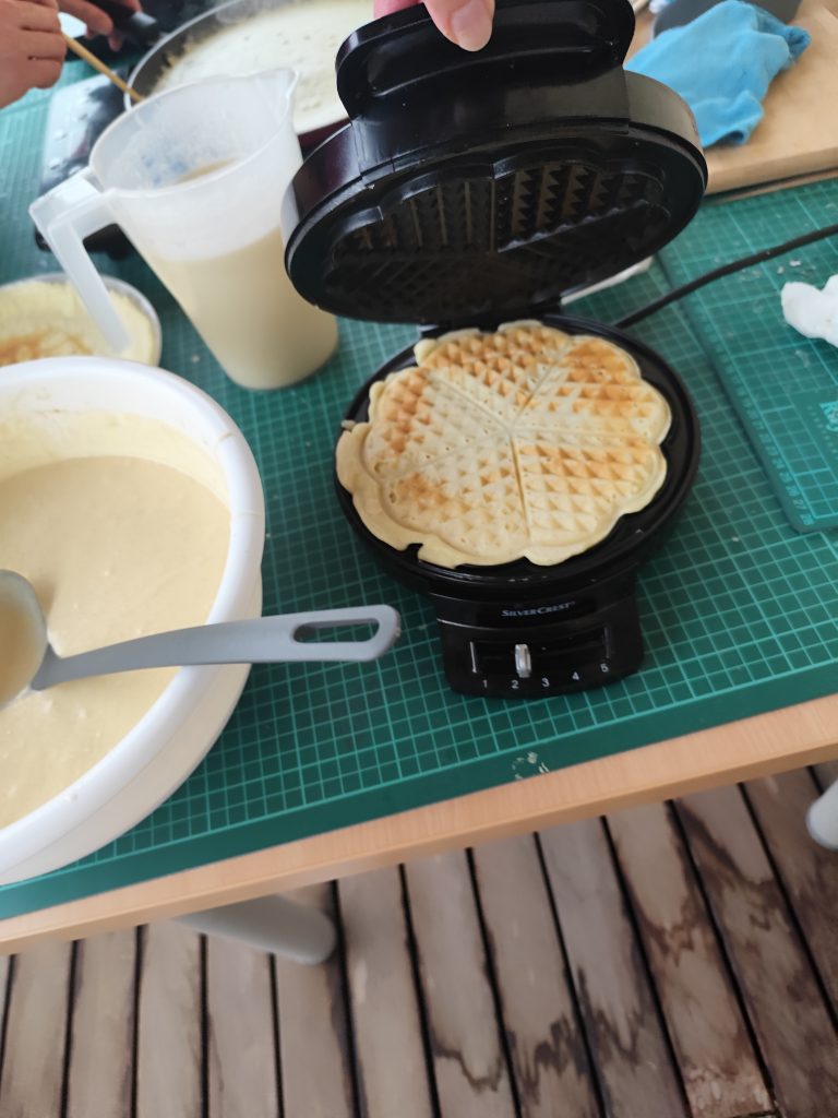 Slika prikazuje pekač v katerem se pripravljajo vaflji, zraven je večja posoda s maso za palačinke.