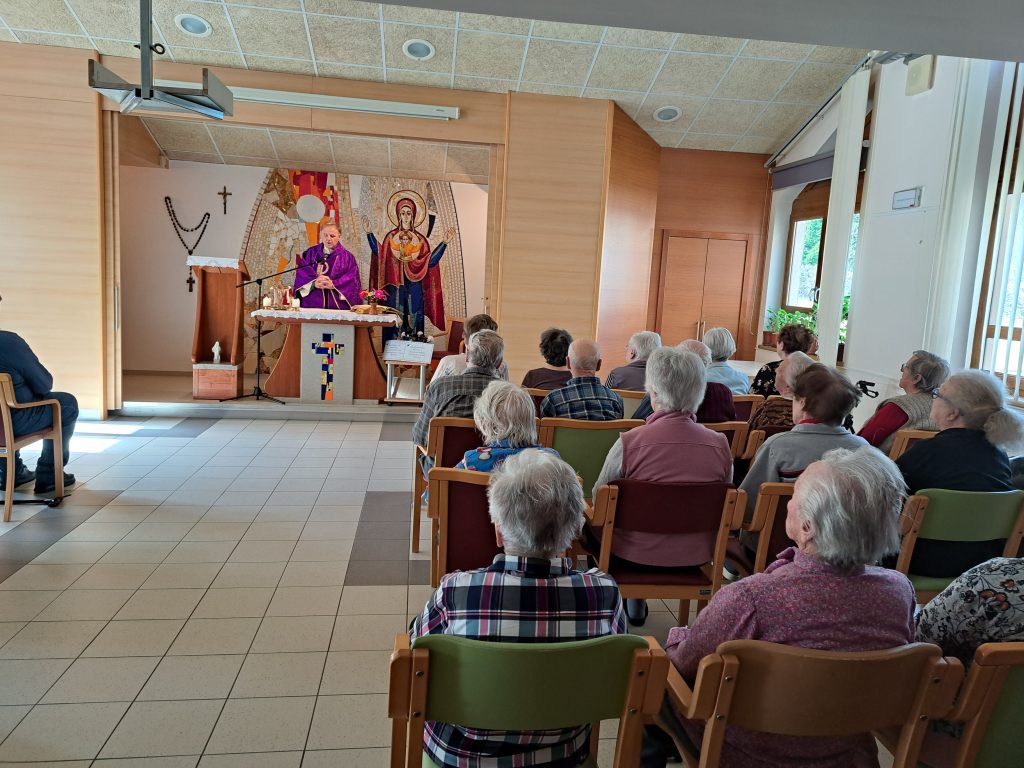 Na sliki je skupina starejših oseb pri mašnem obredu.