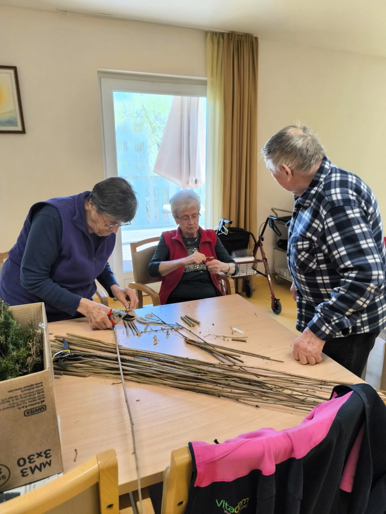 Slika prikazuje tri stanovalce, ki izdelujejo palice iz lesa za nadaljnje izdelovanje velikonočnih butaric.