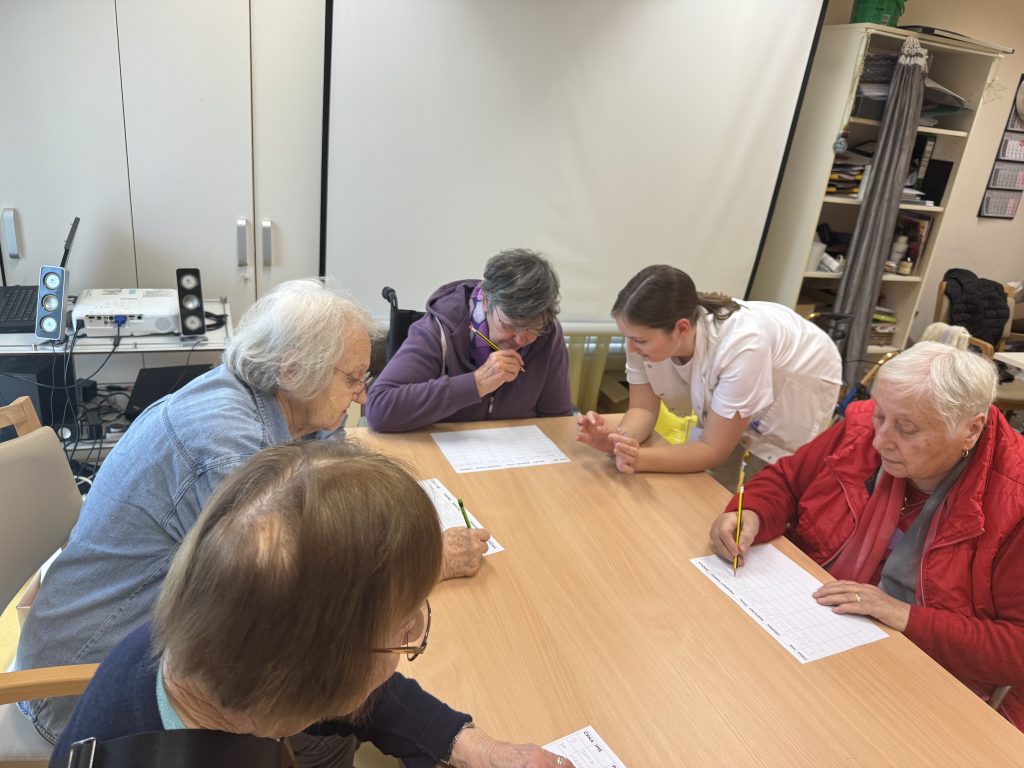 Slika prikazuje stanovalce, ki sedijo pri mizah in so zatopljeni v reševanje nalog na listu papirja. V asistenco jim je študentka delovne terapije.