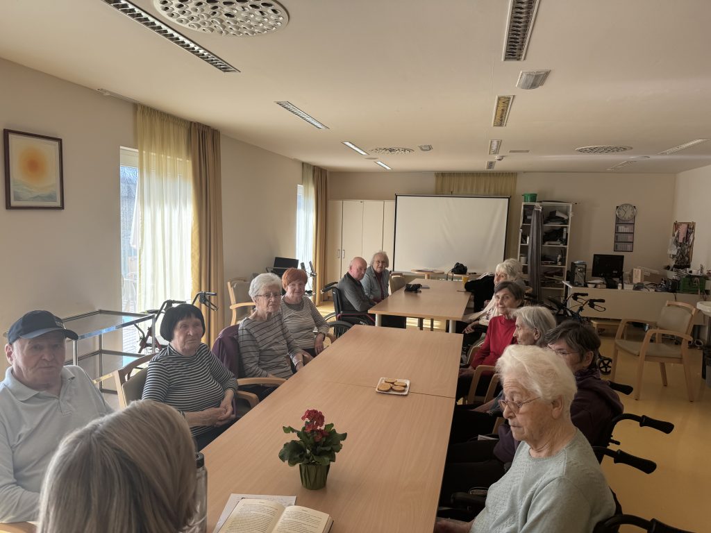 Slika prikazuje stanovalce, ki sedijo na stolih ali vozičkih pri mizi.