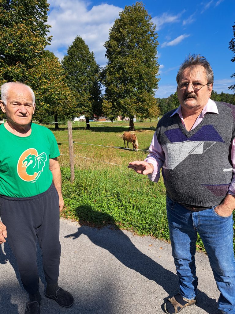 Na sliki sta dve starejši osebi v naravi. V ozadju je konj.