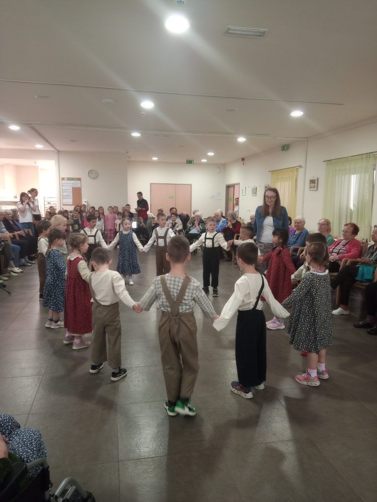Na sliki so v večjem prostoru na sredini v krogu majhni otroci, ki v krogu plešejo. Okrog njih na stolih sedijo starejši.