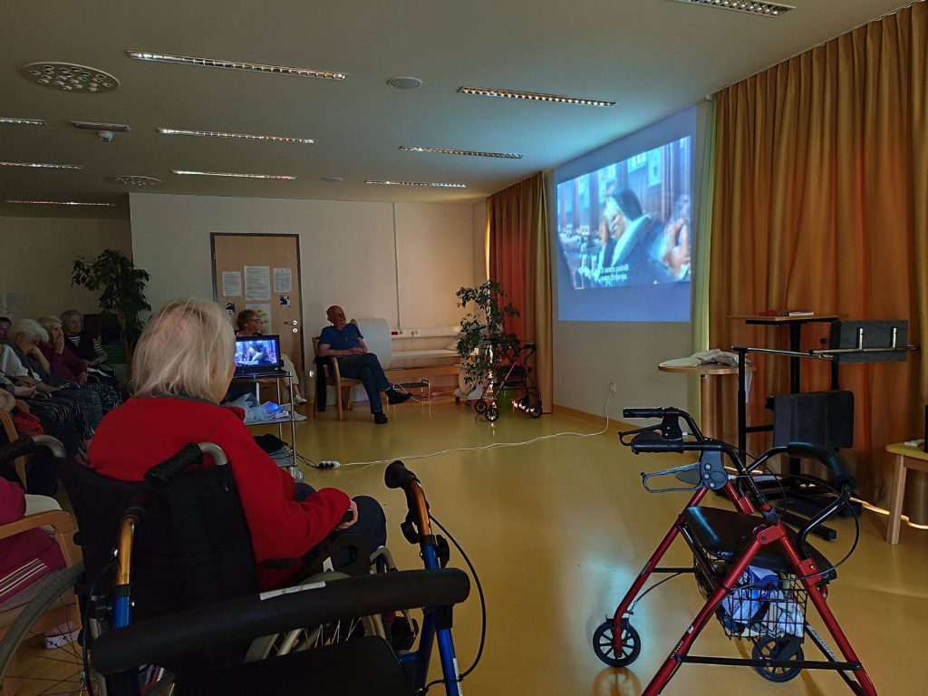 Slika prikazuje sobo v kateri se na platnu predvaja film, stanovalci pa sedijo v polkrogu pred njim.