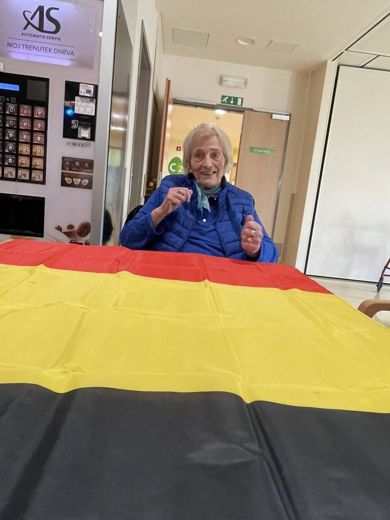 Pri mizi sedi starejša gospa, pred njo je na mizi razprostreta velika belgijska zastava,
