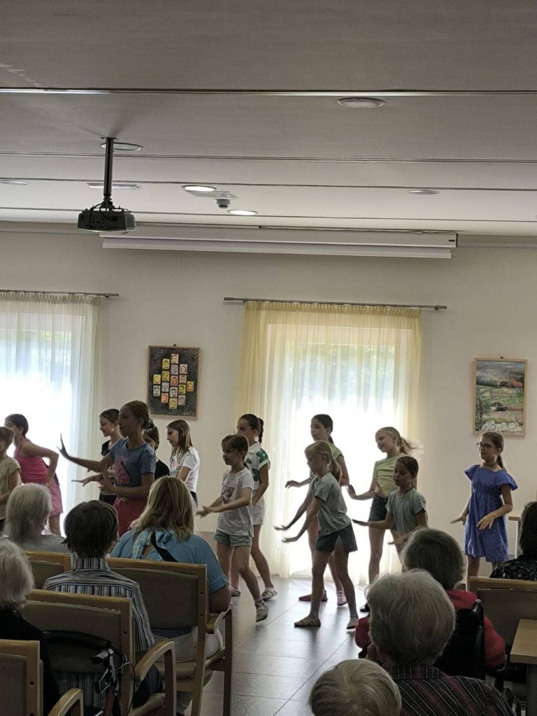 Slika prikazuje skupino osnovnošolk, ki nastopajo pred starejšimi osebami s plesno točko.
