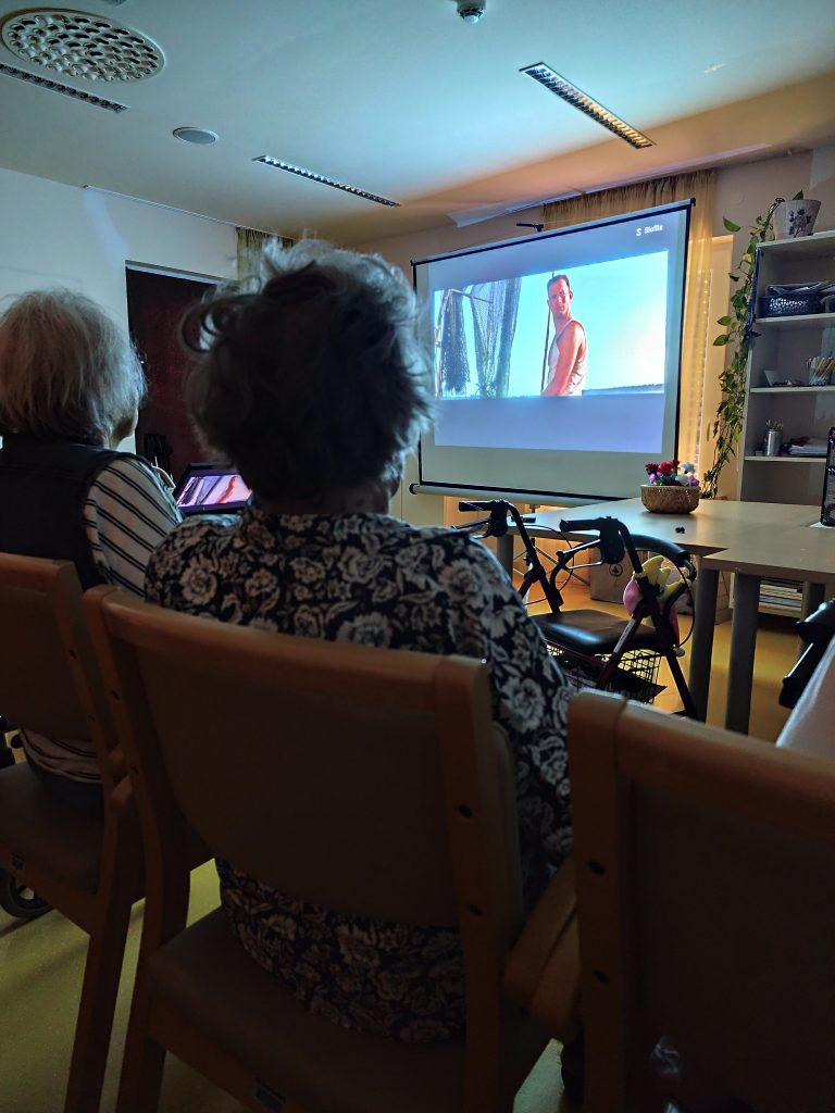 Slika prikazuje platno na katerem se predvaja film, v ozadju pa sedijo stanovalci na stolih.