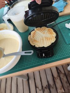 Slika prikazuje pekač v katerem se pripravljajo vaflji, zraven je večja posoda s maso za palačinke.