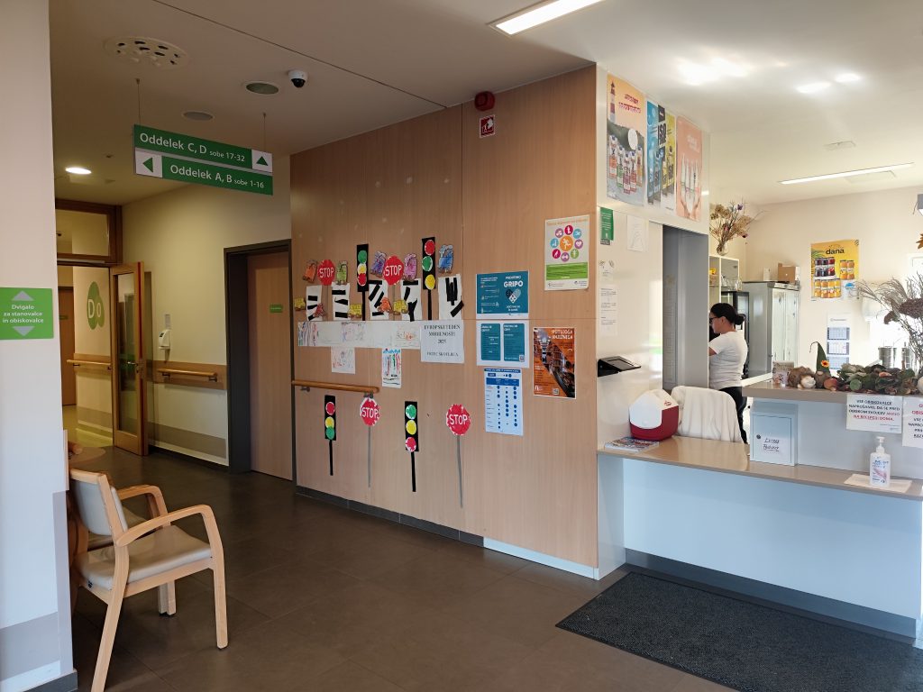 Slika prikazuje vhod v DSO Škofljica, kjer je ob recepciji na steni razstava na temo tedna mobilnosti, vidimo semaforje , STOP znake, avte in prehode za pešce iz papirja.