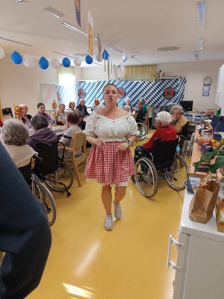 Slika prikazuje vzdušje Oktoberfesta lesene mize, bavarske barve modro-bele v ozadju, na mizah so obešene preste na stojalih, stanovalci pa imajo pred sabo vrčke piva. V ospredju je natakarica v bavarskih oblačilih.