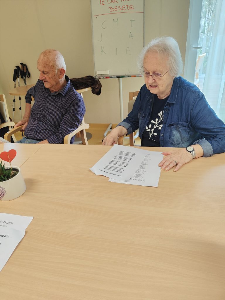 Slika prikazuje stanovalko in stanovalca, ki sedita pri mizi, pred sabo imata list z besedilom, videti je, da pojeta.