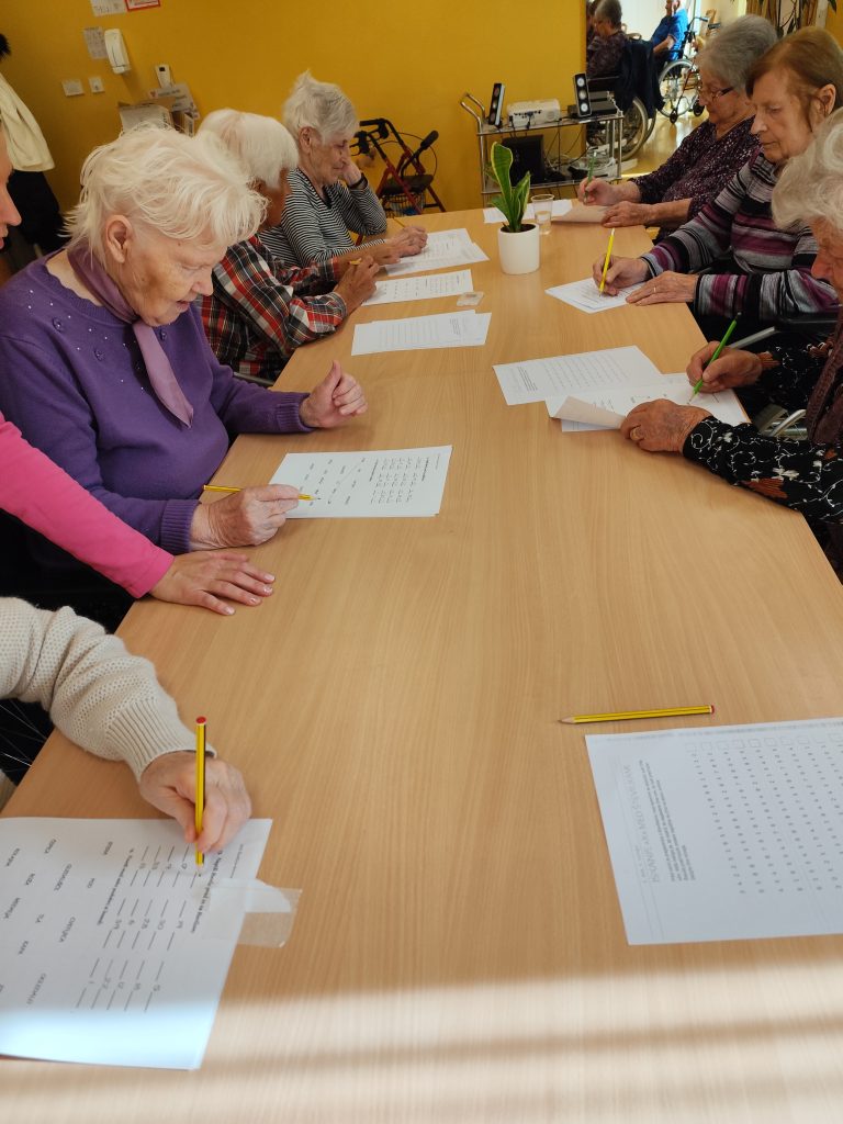 Slika prikazuje skupino stanovalcev, ki ob mizi sedijo in na papirju rešujejo kognitivne naloge.
