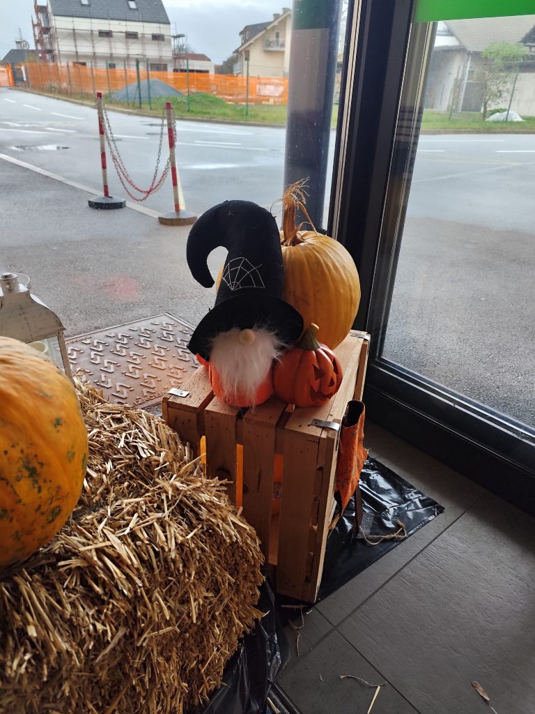 Slika prikazuje malega škrata oranžne barve, z belo brado in črnim klobukom, ki obdan z bučami sedi na lesenem zaboju.