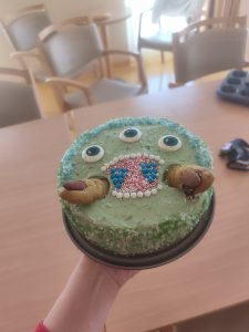 Na slik je zelena okrogla torta s tremi očmi, prsti in odprtimi ustmi.