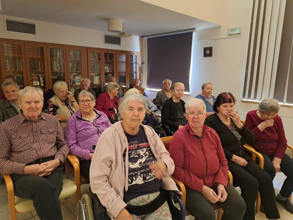 Na sliki je skupina starejših oseb. Sedijo in spremljajo dogojanje.