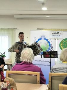 Slika prikazuje mlajšega harmonikaša, ki stoji pred stanovalci DSO, ki sedijo na stolih in so slikani v hrbet