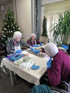 Slika prikazuje 3 gospe, ki sedijo pri mizi in na pekače dajejo kroglice medenjakov.