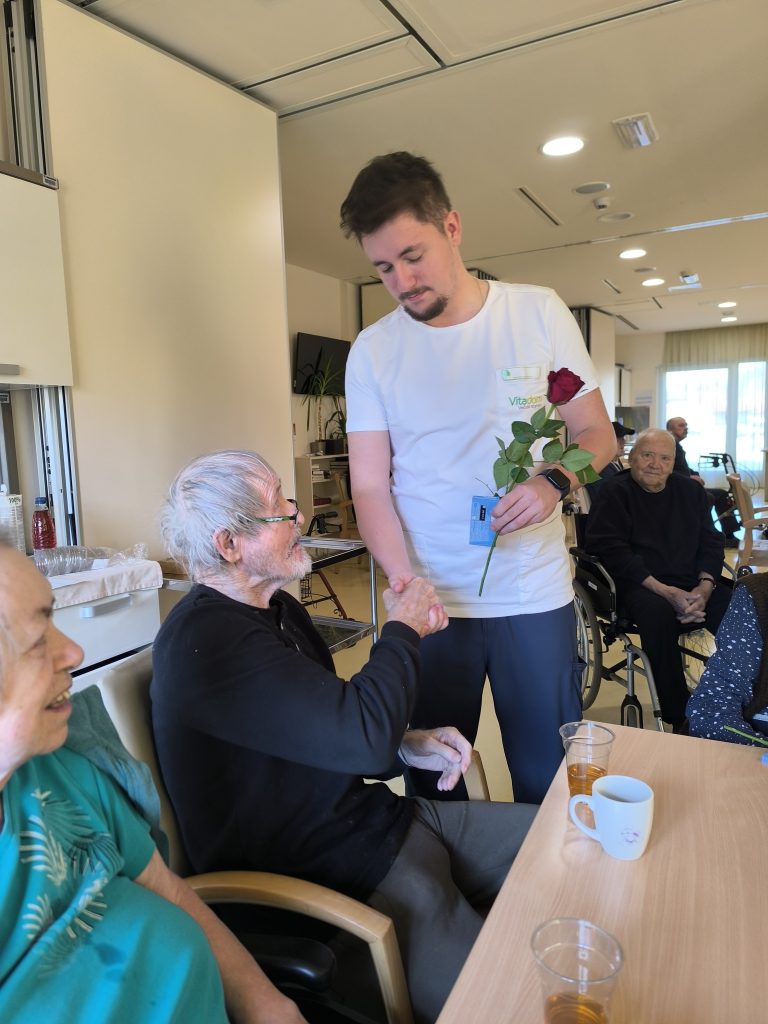 Slika prikazuje zaposlenega, ki stanovalcu ob rojstnem dnevu vošči in mu predaja rdečo vrtnico.